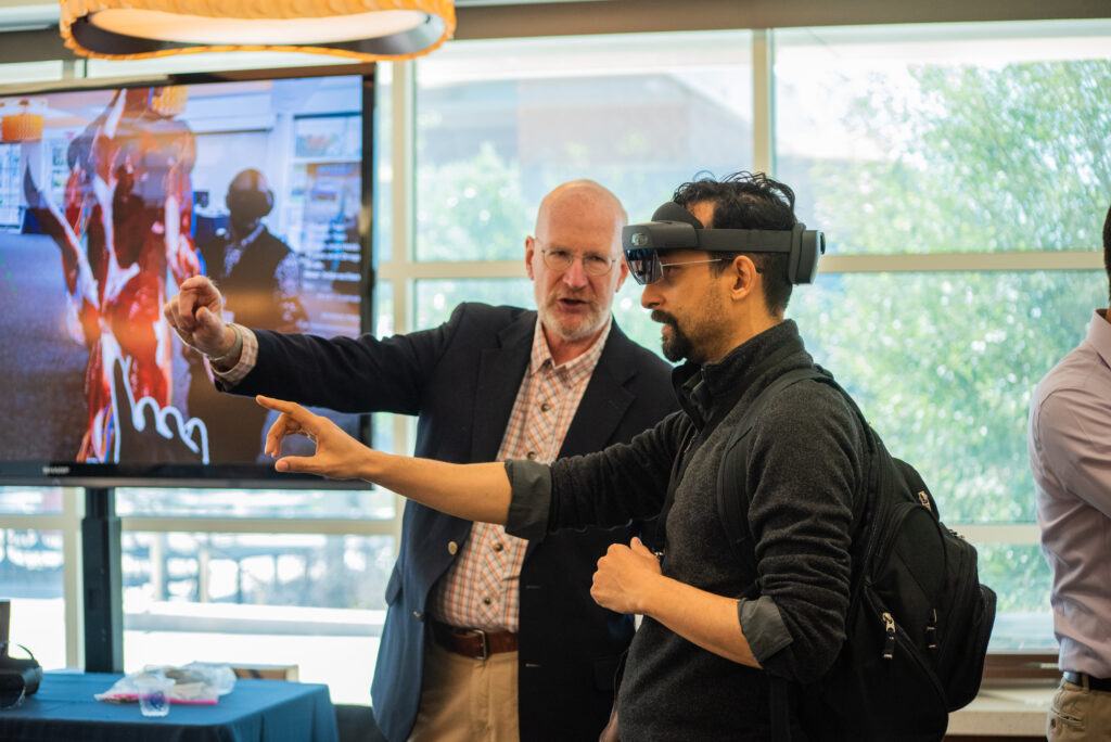 A photo of two individuals, pointing in the same direction. The individual in the foreground is wearing a virtual reality headset.