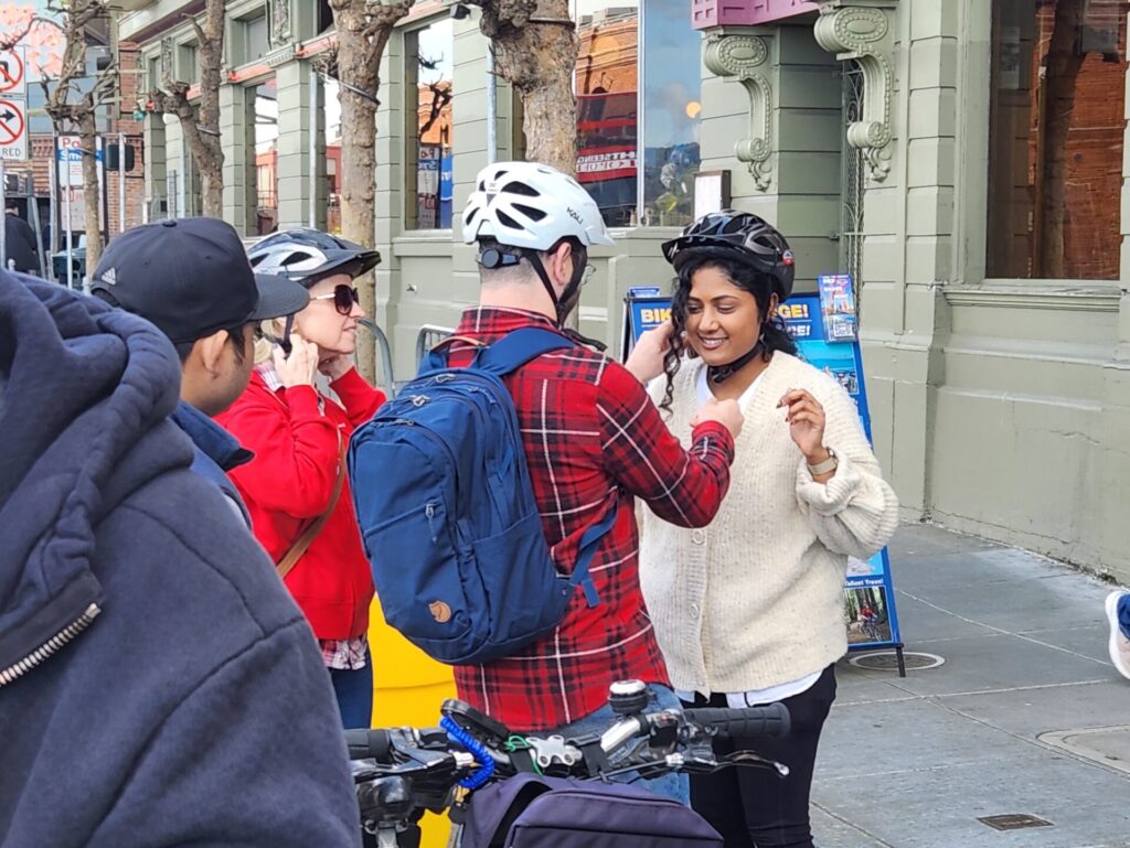 A photo of three individuals wearing bicycle helmets. The individual at center is helping the individual at right with her bicycle helmet.