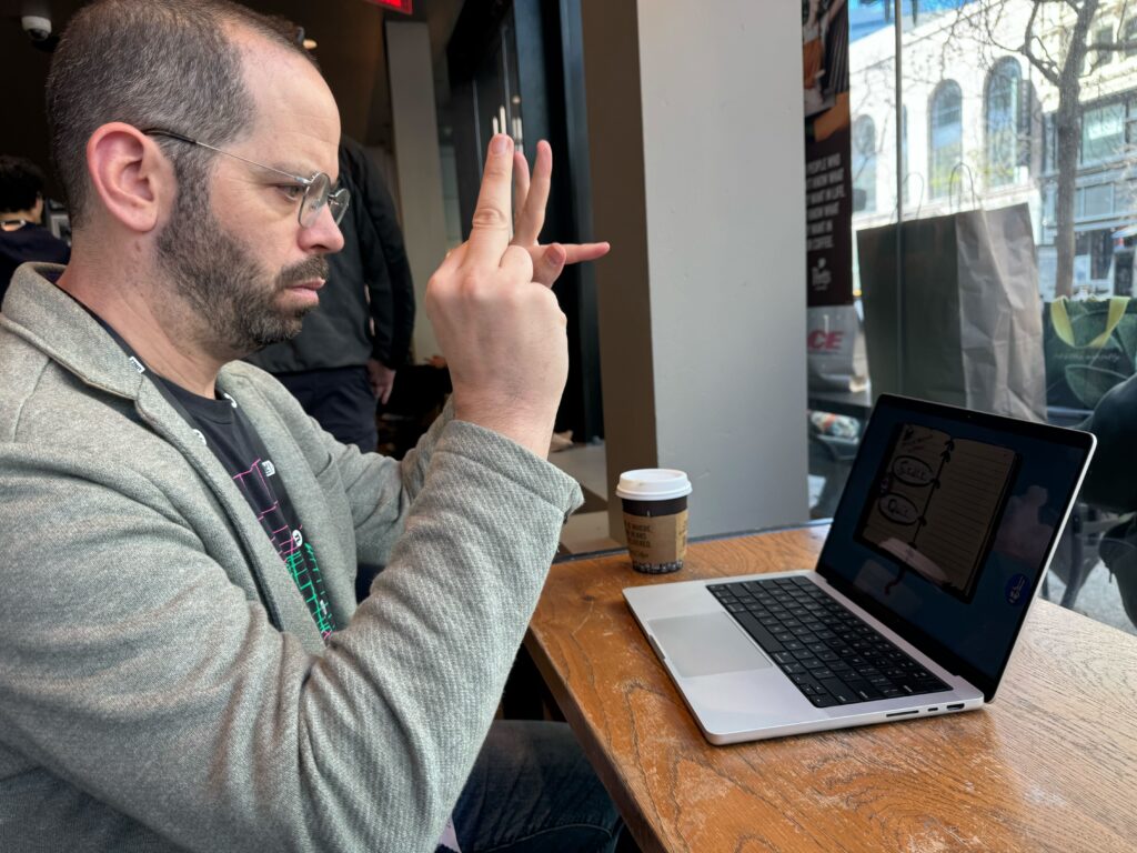 A side view of a person holding a camera up to aa window with a laptop on a table in front of him.
