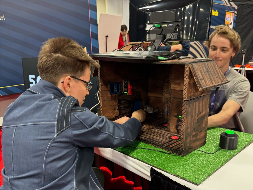 In foreground, an individual in a blazer with short hair reaches into a toy building to move an object. A second individual is seated across from the first on the other side of the toy building.