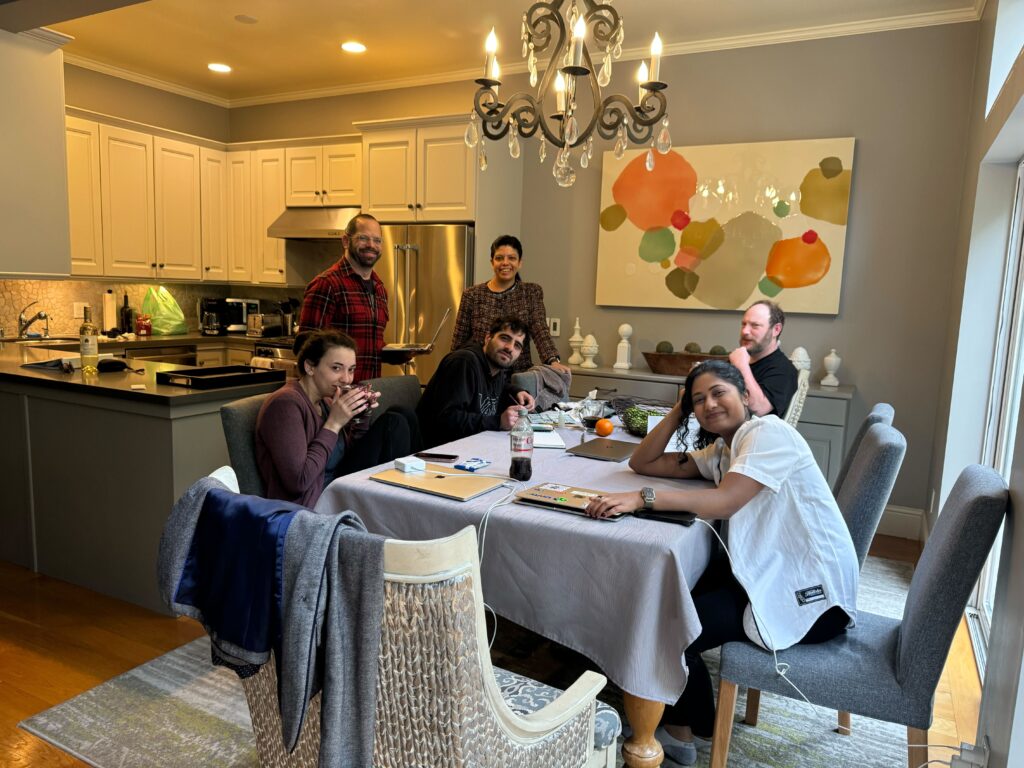 Six people around a kitchen table, with the two in the rear standing. All are looking at the camera.