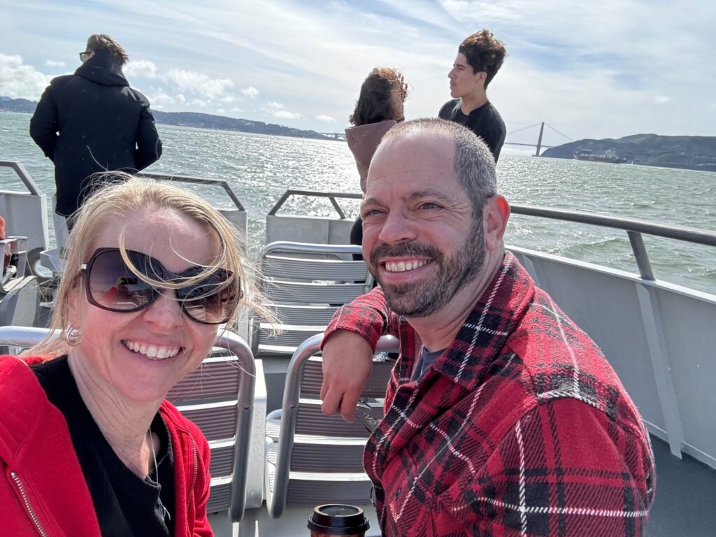 Two smiling people with a boat railing, others, and the San Francisco Bay in the background.