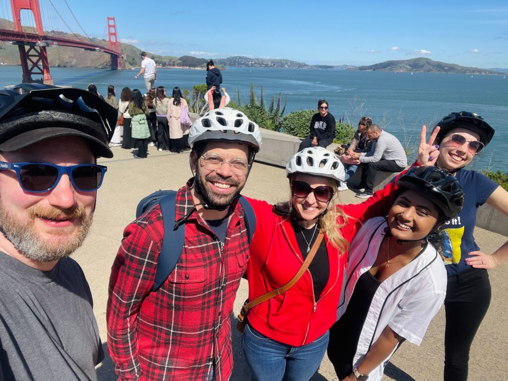 Smiling people in bicycle helmets with the Golden Gate Bridge in the background.