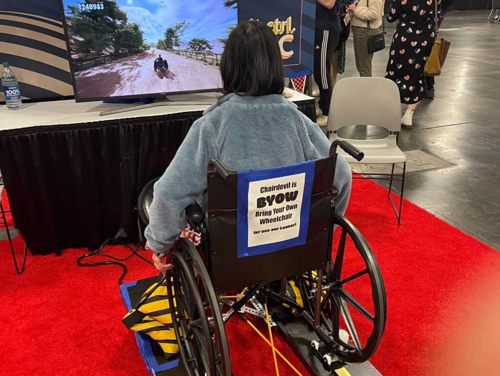 A rear photo of an individual playing a game using a wheelchair and controller that reads wheel motion as input.