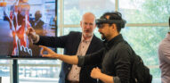 A photo of two individuals, pointing in the same direction. The individual in the foreground is wearing a virtual reality headset.